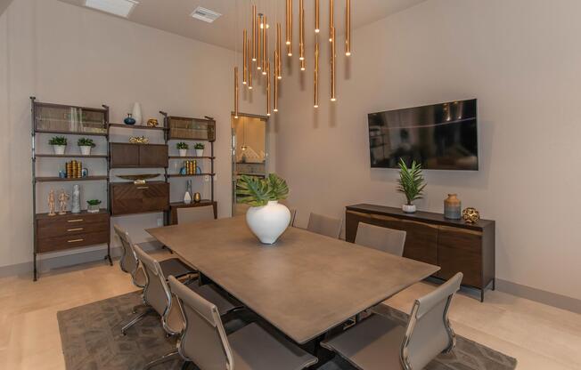 A modern conference room featuring a large rectangular table surrounded by sleek chairs. Above, there are contemporary pendant lights hanging from the ceiling. A flat-screen TV is mounted on the wall, and decorative shelves display plants and accessories. The room has a neutral color palette with soft lighting.