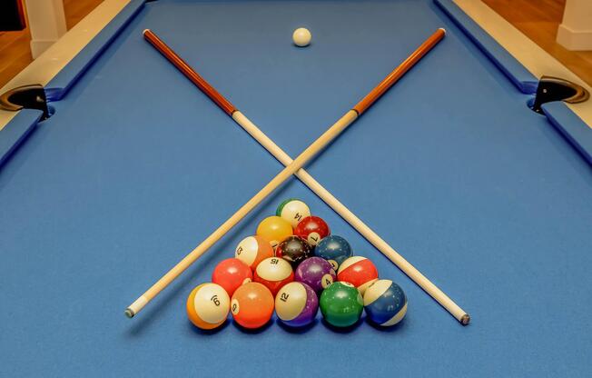 A billiard table featuring a set of colorful pool balls arranged in a triangle formation at the center. Two cue sticks are crossed over the triangle, with a white cue ball positioned above it. The table is covered in blue felt, creating a vibrant playing surface.