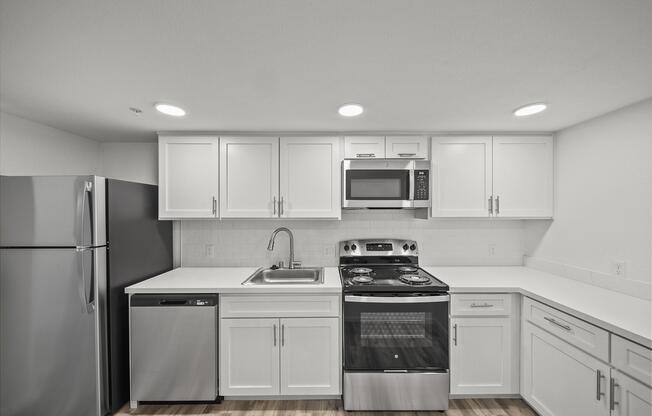 Modern kitchen featuring white cabinetry, a stainless steel refrigerator, a silver dishwasher, and a gas stove with an oven. The kitchen also includes a microwave and a stainless steel sink on a white countertop, illuminated by recessed lighting. Wood laminate flooring completes the contemporary look.
