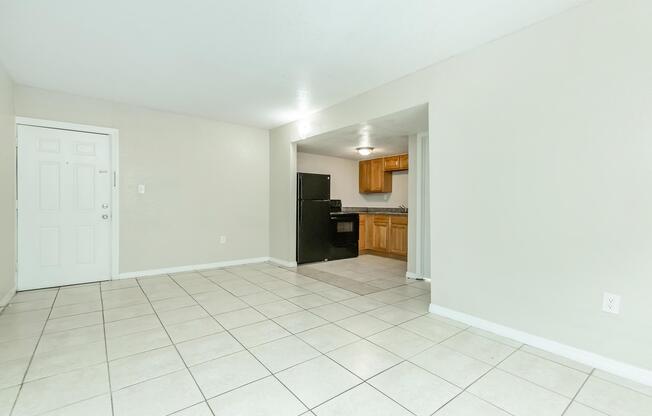 Interior view of a spacious, light-colored living area with tile flooring. To the left, a white door leads outside. In the background, a kitchen with wooden cabinets and black appliances is visible through an open entryway. The space is clean and bright, showcasing a simple and modern design.