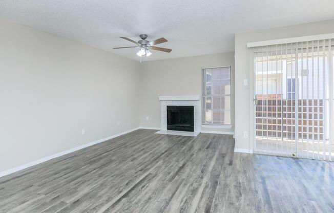A spacious living room with light-colored walls and wood-look laminate flooring. It features a ceiling fan, a fireplace with a white mantel, and large windows with blinds. A sliding glass door leads to a balcony or patio area. The overall design is modern and inviting, with plenty of natural light.