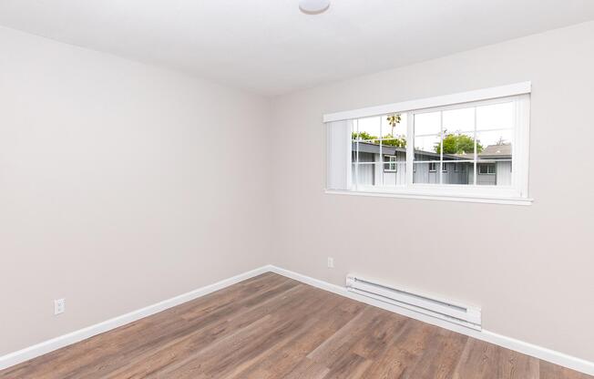 Empty room with light beige walls and large window. The floor is covered with hardwood-looking laminate. A baseboard heater runs along one wall. Natural light enters through the window, highlighting the spaciousness. The overall atmosphere is clean and minimalistic.