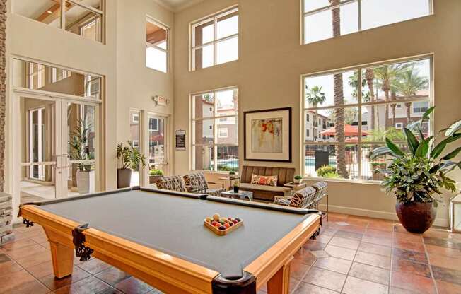 A vibrant clubhouse game room here at Mozaic at Steele Park featuring a full-size billiards table set on tile flooring beneath soaring ceilings and expansive windows. Comfortable lounge seating and lush potted plants create a relaxed atmosphere, while large windows showcase views of the palm-lined pool courtyard just outside, filling the space with abundant natural light.