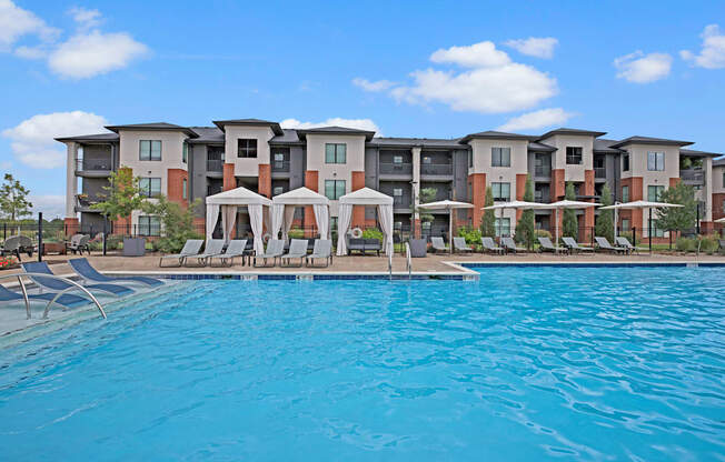 Luxurious poolside view of 49 West's modern apartment complex, featuring cabanas and lounge chairs under a blue sky with fluffy clouds. Relaxing and inviting atmosphere.