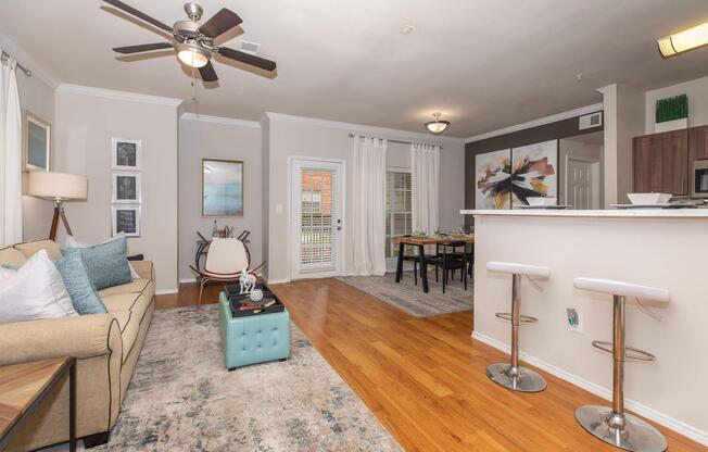 A cozy living area featuring a beige sofa with decorative pillows, a turquoise coffee table, and a stylish rug. Thereâs a ceiling fan above, a dining area with a wooden table, and modern bar stools. Large windows allow natural light, and artwork adorns the walls, creating a welcoming atmosphere.