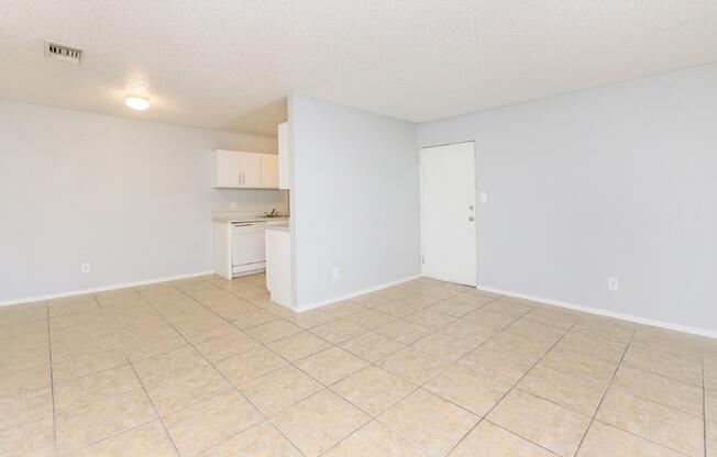 A spacious, empty room featuring light-colored tiled flooring, beige walls, and a simple kitchen area with white cabinets in the background. A door leading outside is visible on the right, and a ceiling light fixture illuminates the space, creating a bright and open atmosphere.