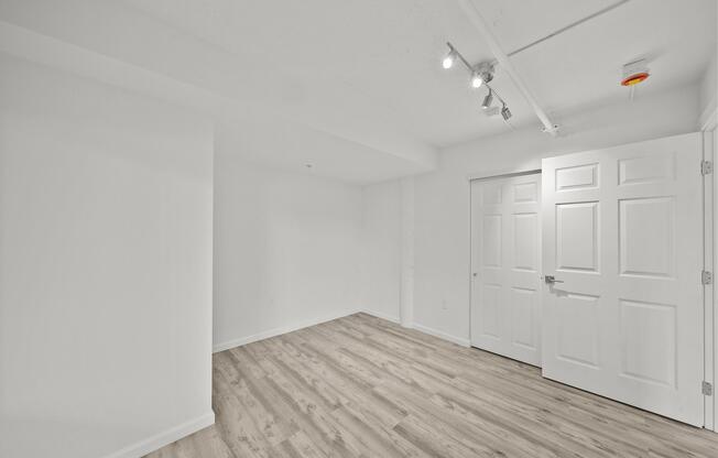 A spacious, empty room with white walls and a light wooden floor. Two doors are visible: one is a double door leading to a closet, while the other is a standard door. The ceiling features track lighting and there is a small overhead light. The room is well-lit and has a clean, modern aesthetic.
