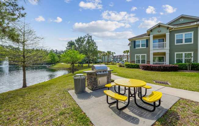 A yellow picnic table is in the foreground of a park with a lake and apartment building in the background.