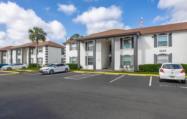 A residential apartment complex featuring two buildings with white exteriors and brown roofs. There are several parked cars in the foreground and a staircase leading to an entrance. A flag is visible on one building, with partly cloudy blue skies above.