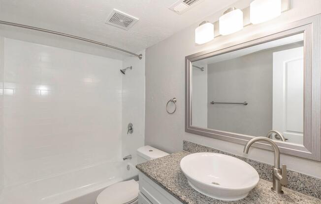 A modern bathroom featuring a white bathtub with a shower, a granite countertop with a round sink, a wall-mounted mirror, and three light fixtures above. The walls are painted in light gray, with a towel rack and a white door visible in the background.