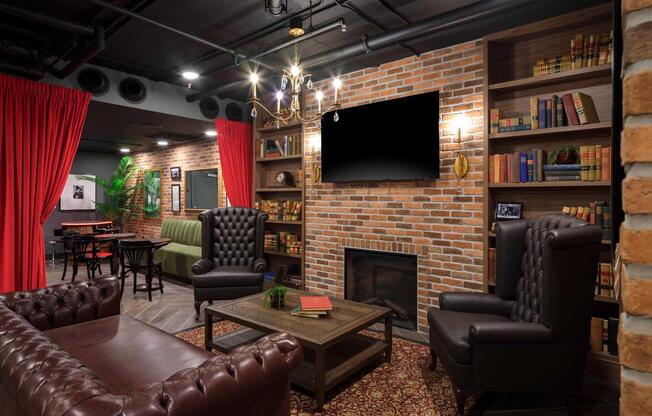 Cozy lounge area featuring a brick wall, dark wood bookshelves filled with books, two leather armchairs, a coffee table with decorative items, a fireplace, and a large television mounted on the wall. Red curtains frame the space, adding warmth to the atmosphere. A green upholstered sofa is visible in the background.