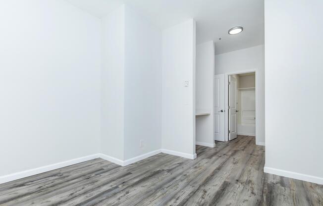 A bright, empty room with light-colored walls and a wood-like floor. There is a doorway on the right leading to a bathroom, and a small closet space visible. The ceiling has a round light fixture, and the overall atmosphere is clean and modern, suitable for various uses or furnishings.