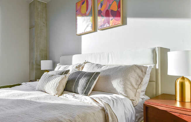 A bed with white pillows and a white comforter is in the foreground of a room with a painting on the wall.