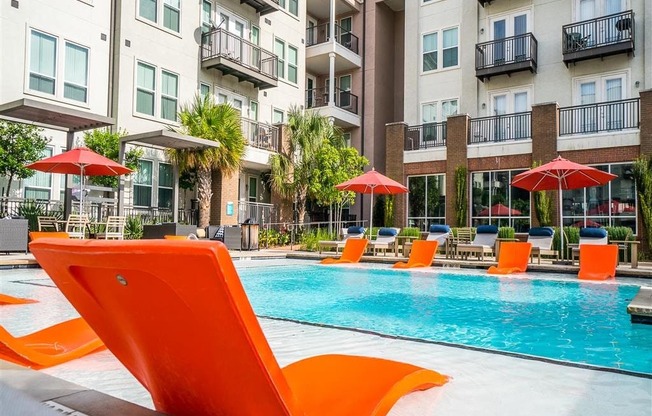 A modern pool courtyard here at BRIO at Firewheel with a clear swimming pool, bright orange in-water lounge chairs, shaded seating with umbrellas, and surrounding apartment balconies and landscaping.