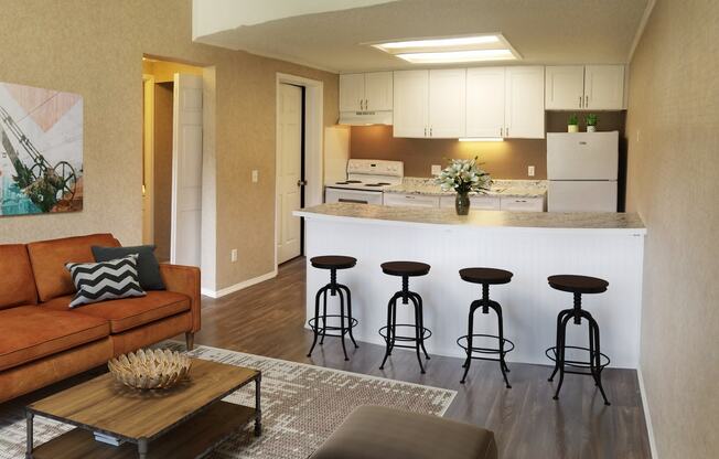 A kitchen with white cabinets and a refrigerator is adjacent to a living room with a brown couch and a coffee table.