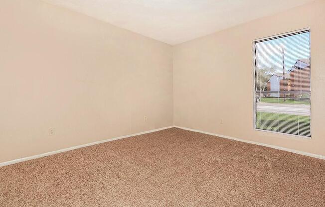 A vacant room with beige walls and a light brown carpet. There is a window with blinds on one wall, allowing natural light to enter. The space is empty, giving it a spacious feel, and there are no furnishings or decorations.
