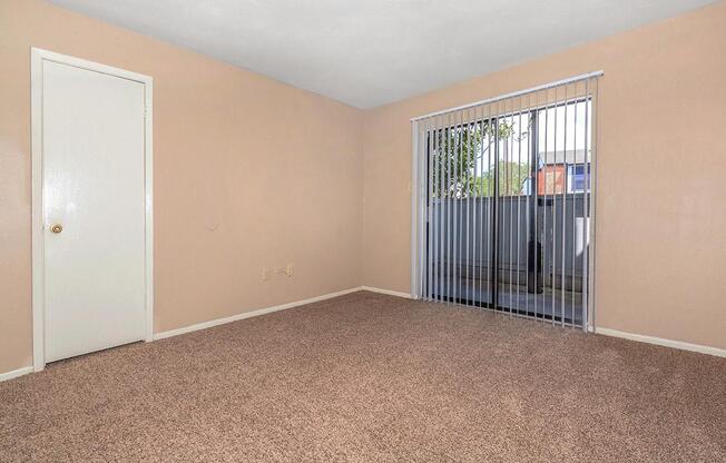 Empty room with light beige walls and carpeted floor. There is a closed white door on the left and a large window with vertical blinds that lets in natural light. Outside the window, a fence is visible. The room appears spacious and ready for furniture.