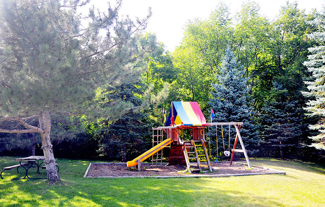outdoor playground with slide and swing next to a park bench and large trees all around