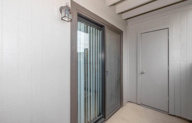 A view of a modern entrance area featuring a sliding glass door with vertical blinds, a framed solid door, and a small lantern on the wall. The walls are painted in a light color with a textured finish, and the ceiling has exposed beams, creating a bright and inviting atmosphere.