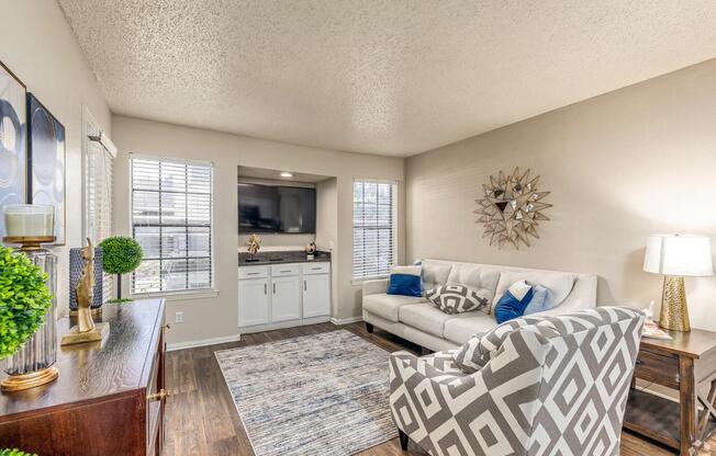 A cozy living room featuring a light-colored sofa with blue accent pillows, a patterned armchair, a small bar area with a flat-screen TV, and decorative items. Natural light filters through the window blinds, highlighting a stylish rug and a decorative mirror on the wall.