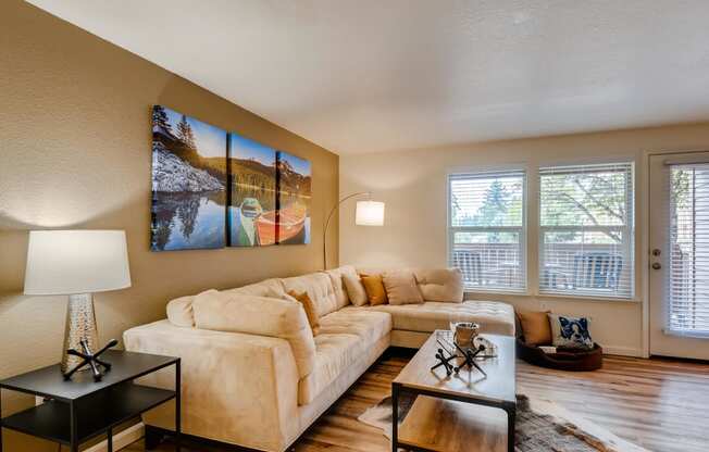 Apartments Near Happy Valley For Rent - Sunnyside Village - a Living Room with Wood-Style Flooring, a Large Plush Couch, a Coffee Table, a Small Table with a Lamp, Large Windows with White Blinds, and a Door to the Private Patio