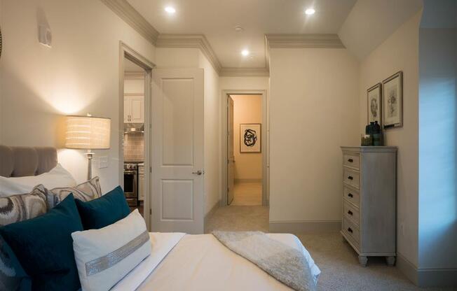 A cozy bedroom with navy and cream bedding, a dresser, and a hallway to the kitchen.