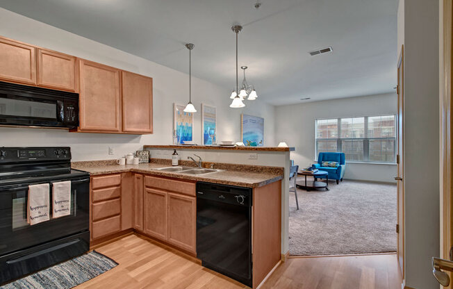 A bright kitchen and living area here at Swan Creek with warm wood cabinets, black appliances, and open breakfast bar. Cozy living room beyond features soft carpet and a standout teal accent chair.