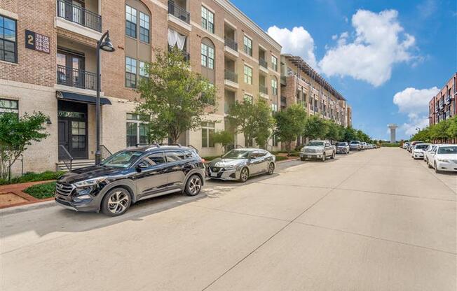 A black car is parked on the left side of a street lined with parked cars and apartment buildings.