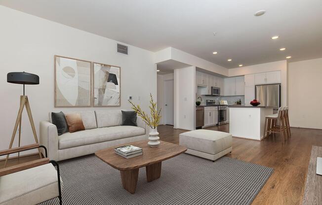 Modern living room and kitchen area featuring a light-colored sofa with decorative pillows, a wooden coffee table, and a patterned rug. The kitchen is equipped with stainless steel appliances and white cabinets. Natural light fills the space, complemented by wall art and a floor lamp.