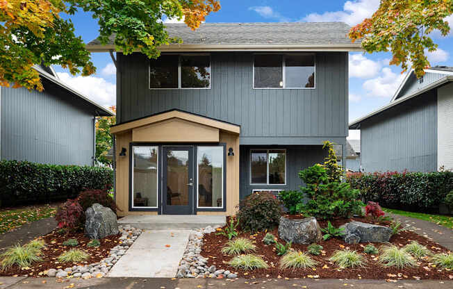 A house with a grey exterior and a brown door is surrounded by a garden with red and green plants.