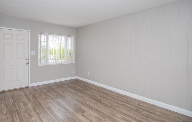 A light gray living room with wood-like laminate flooring. The space features a white door on the left and a window with blinds allowing natural light in. The walls are painted light gray, and the area is uncluttered, creating a simple and inviting atmosphere.