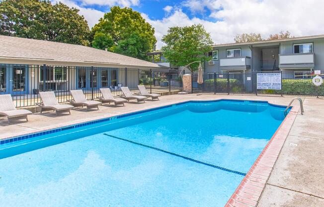 A clean and inviting swimming pool surrounded by lounge chairs, with a well-maintained courtyard and buildings in the background. The sky is partly cloudy, and there are trees providing a pleasant atmosphere. Ideal for relaxation and leisure activities.