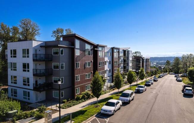 Drone shot of the apartment building showcasing the exterior of the building and the neighborhood. Cars parked along the street  in front of the building and clear blue skies outside.