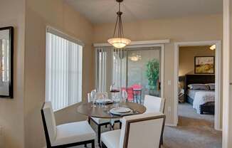 A bright dining area here at Dobson Towne Center featuring plush carpet, a round dining table with seating for four, and a modern pendant light overhead. Large windows and a sliding glass door provide natural light and access to a private patio, while an open doorway leads to a spacious bedroom, creating a connected and comfortable layout.