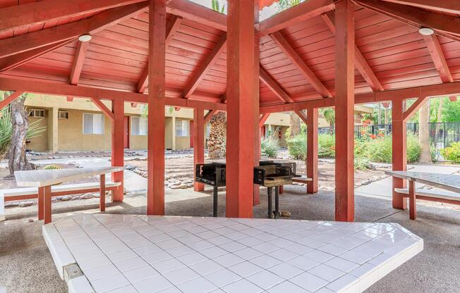 An outdoor pavilion with a red wooden structure featuring a central grilling area and several white tile-topped tables. Surrounding the pavilion are small palm trees and landscaped areas. The background shows a residential building with beige walls.