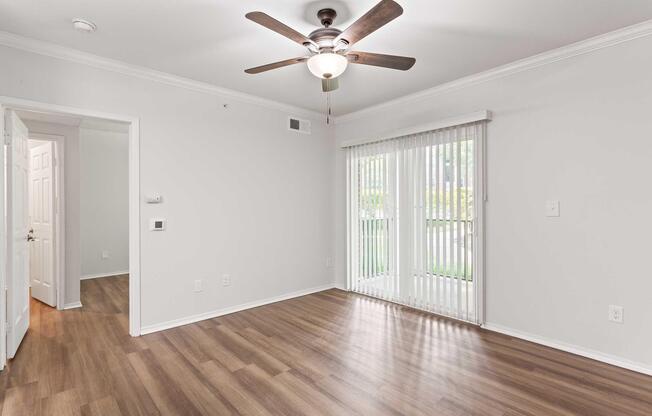 A well-lit room with light gray walls, hardwood flooring, and a ceiling fan. A sliding glass door with vertical blinds allows natural light. An entrance leads to another room, while a wall features a small control panel. The space appears clean and minimally decorated.