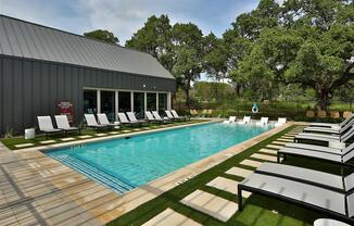 A refreshing swimming pool surrounded by green space with patios, lounge chairs, and large trees.