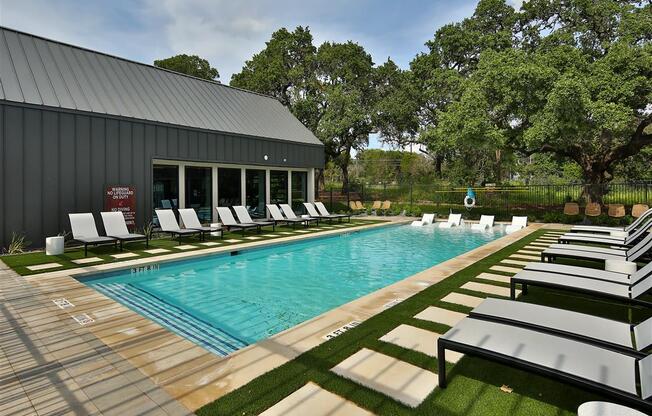 A refreshing swimming pool surrounded by green space with patios, lounge chairs, and large trees.