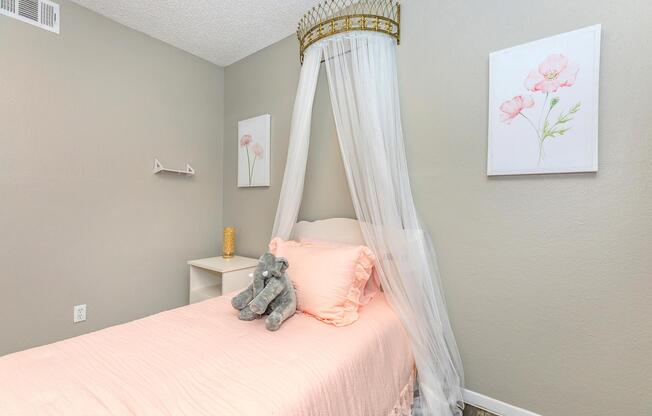 A softly decorated bedroom featuring a single bed with a light pink duvet and decorative pillows. Above the bed, a whimsical canopy hangs from the ceiling, and a plush gray elephant is resting on the bed. The walls are painted gray, adorned with two floral artwork pieces. There's a small nightstand beside the bed.