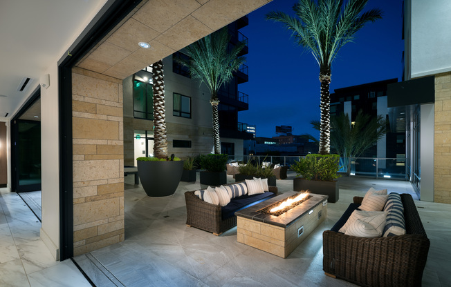 Outdoor firepit and lounge area adjacent to the indoor clubhouse with full-length windows that slide away to create an indoor-outdoor space. There are two wicker couches on opposite sides of the firepit.
