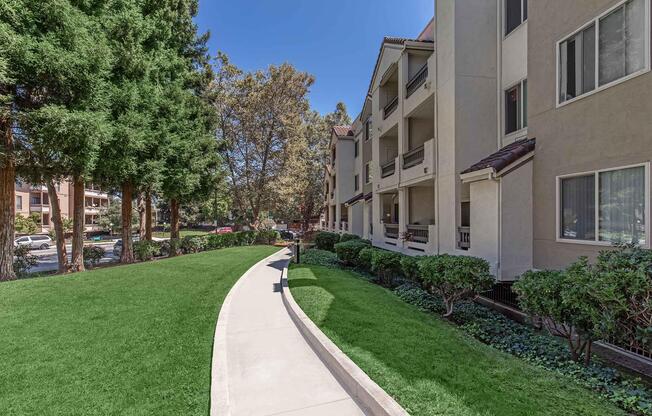 A landscaped pathway curves through a grassy area, flanked by neatly trimmed bushes and trees. In the background, residential apartment buildings are visible, with balconies and windows. The scene is set under a clear blue sky, giving a warm and inviting atmosphere.
