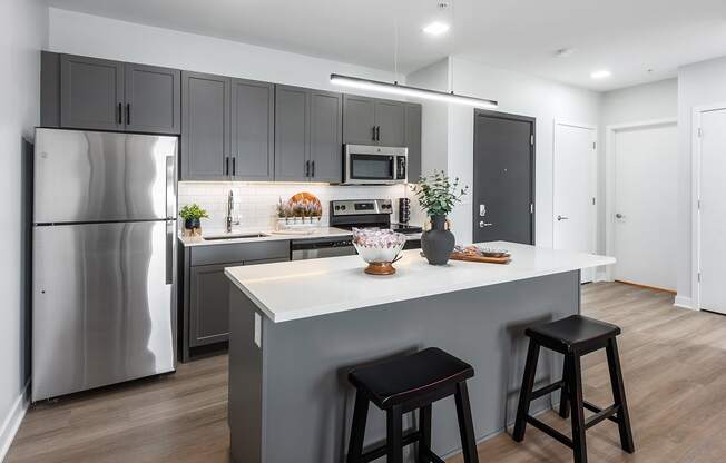 model kitchen with stainless steel appliances, plank flooring, kitchen island, white quartz countertops, gray cabinetry and white tile backsplash at Steelcote Square, St. Louis, 63103