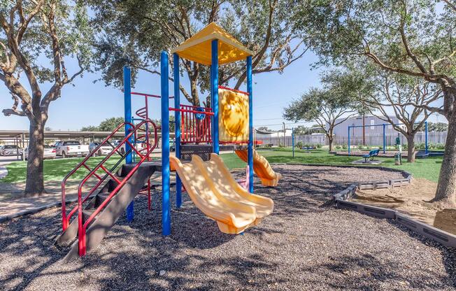 Colorful playground equipment featuring a slide, climbing structure, and play area surrounded by trees. The ground is covered with mulch, and nearby, there are visible vehicles and a grassy area under a clear blue sky. Ideal for children's outdoor play and activities.