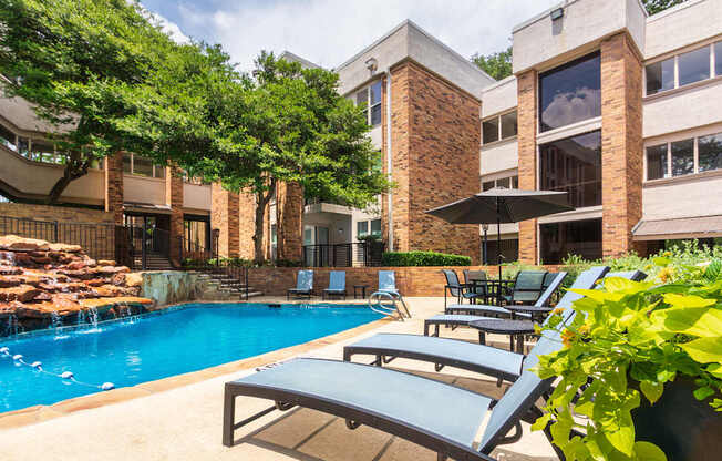 A swimming pool with a waterfall surrounded by lounge chairs and a couple of tables, chairs and umbrellas at Cambridge Court Apartments Entryway in Lake Highlands, Dallas, TX.