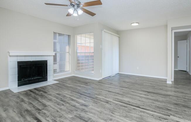 A spacious, well-lit living room featuring a ceiling fan, a modern fireplace with a white surround, large windows with blinds, and a sliding glass door. The flooring is a light-colored laminate, and the walls are painted in a soft neutral tone. There is an adjacent doorway leading to another room.