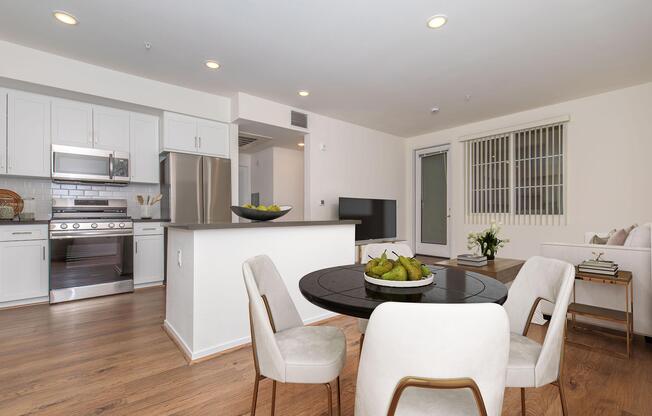 Modern apartment interior featuring a kitchen with white cabinets and stainless steel appliances, a dining area with a round black table and four white chairs, and a living space with a light-colored sofa and TV. Large windows allow natural light, creating a bright and inviting atmosphere.