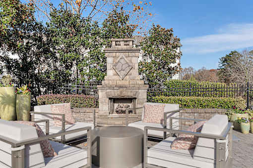 A fireplace is in the middle of a patio with white chairs around it.