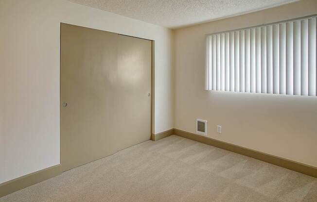 A bright, empty bedroom here at Whispering Cedars with soft carpet, neutral walls, and a window with vertical blinds, featuring a spacious sliding door closet and a clean, comfortable layout.
