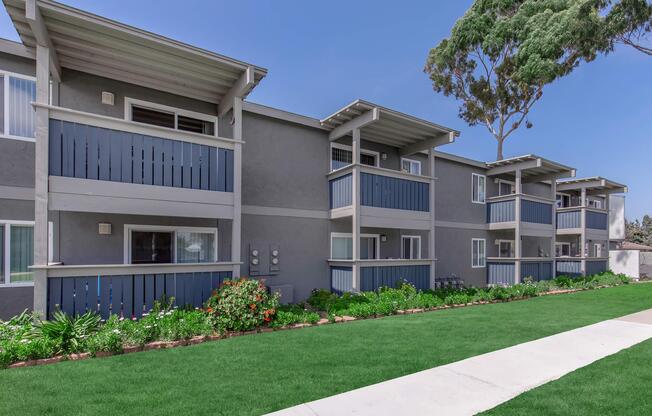 A modern apartment complex featuring multiple units with balconies, surrounded by lush green landscaping and a clear blue sky. The building has a gray exterior with blue accents and well-maintained flower beds along a paved walkway.
