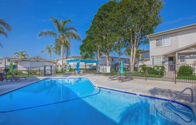 A serene swimming pool surrounded by palm trees and lounge chairs. A shaded gazebo is in the background, with white buildings lining the area. The sky is bright blue, creating a relaxing atmosphere suitable for leisure and outdoor activities.